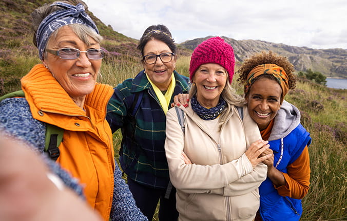 Front view of a small group of mature and senior female friends hiking up the mountain landscape surrounding Loch Torridon in the Northwest Highlands, Scotland. A senior woman is wearing a wooly hat and the rest are wearing bandanas, they are side by side taking a selfie on a smartphone.