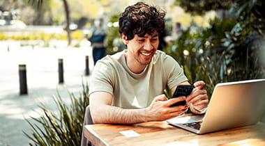 Man smiling and working on laptop in public park, while looking at his cellphone