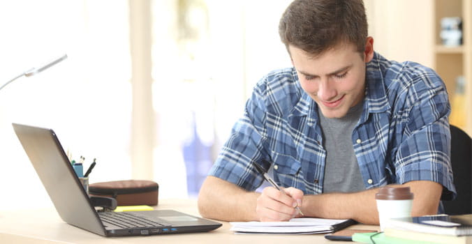 Young man writing in notebook with laptop next to him