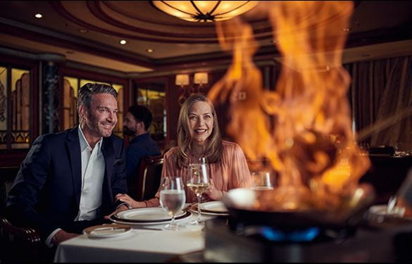 Two diners aboard a Cunard cruise ship, watching a meal prepared at their table with a roaring flame