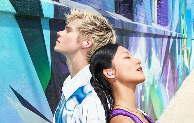 A man and woman stand back to back, both wearing colorful clothing and earphones, amidst a colorful wall mural on a building.