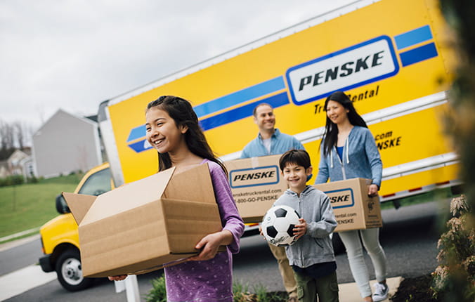 Family of 4 carries moving boxes in front of a Penske rental truck